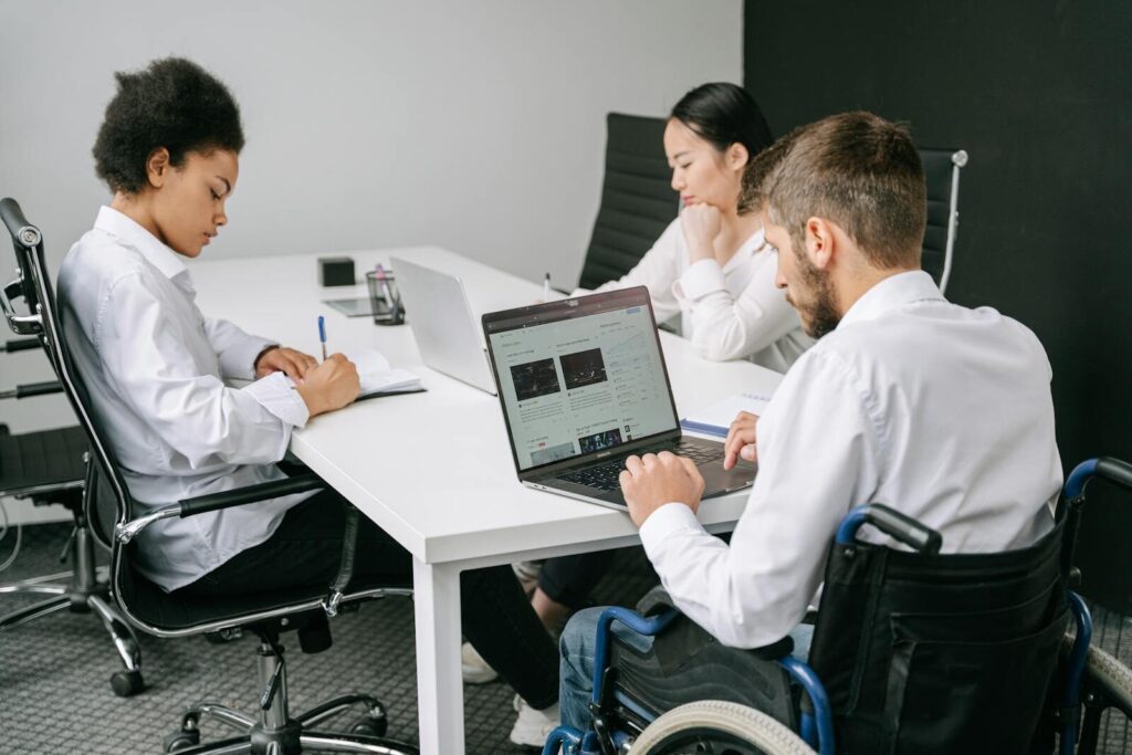 Three diverse colleagues, including a person in a wheelchair, collaborate in an office using laptops and notes.
