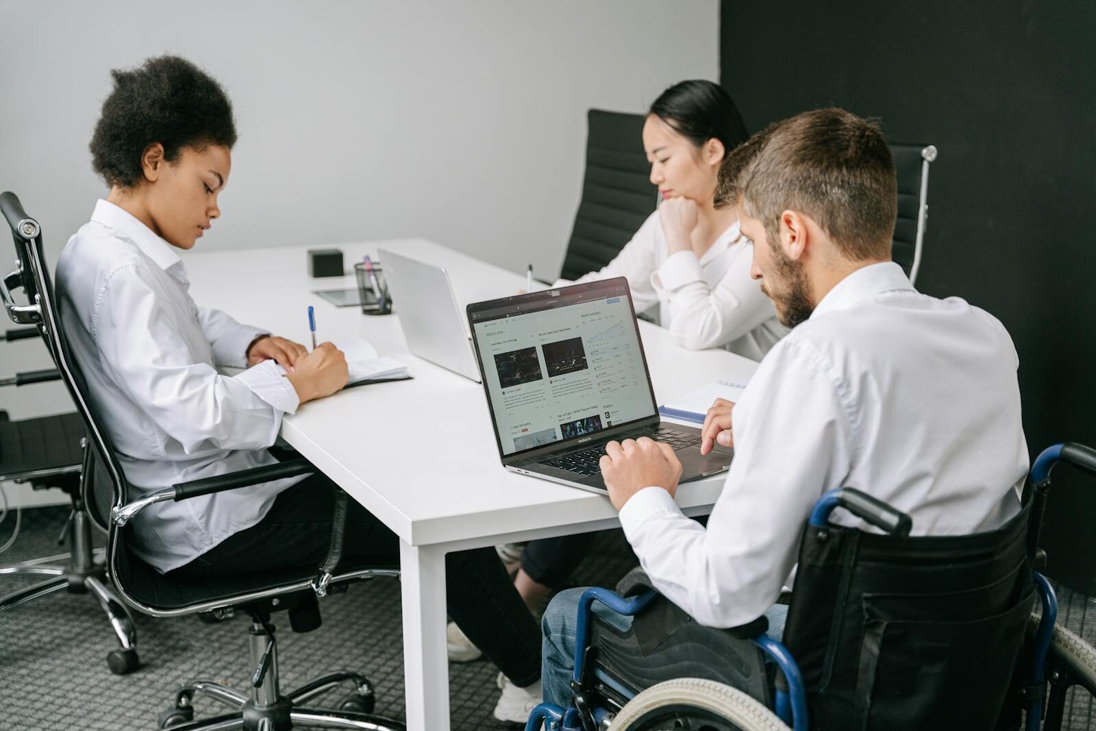 Three diverse colleagues, including a person in a wheelchair, collaborate in an office using laptops and notes.